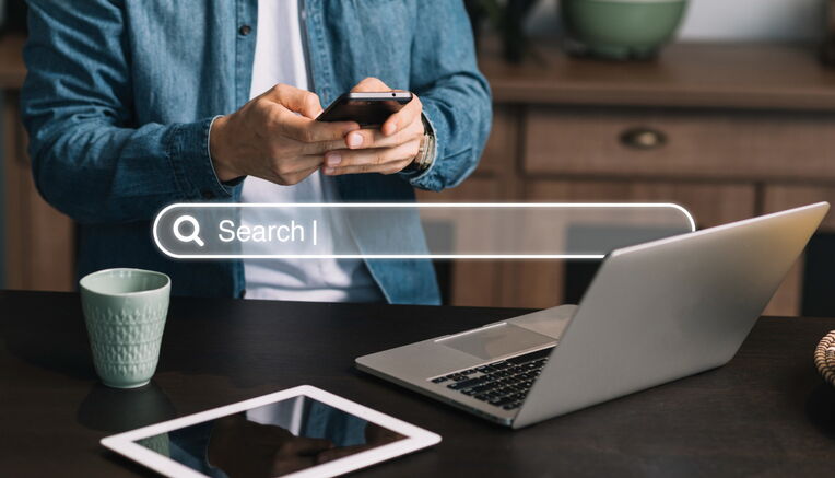 Ein Mann nutzt ein Smartphone vor einem Laptop und Tablet mit einer eingeblendeten Suchleiste. Dies könnte die Recherche nach Keywords für Produktbeschreibungen über verschiedene Geräte symbolisieren.