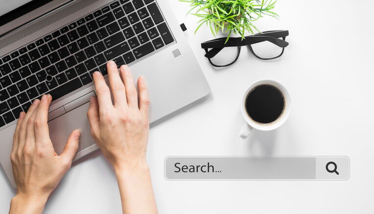 Hände tippen auf Laptop mit einer Suchleiste. Daneben Kaffeetasse, Brille, Pflanze. Visualisiert das "Content mit Keywords aufwerten" für effizientes Arbeiten.